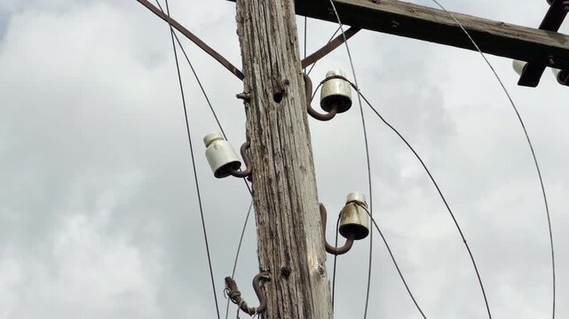 A close up shows white ceramic insulators on a weathered wooden pole holding drooping wires. The scene conveys a lonely and nostalgic mood of forgotten times under a cloudy sky.