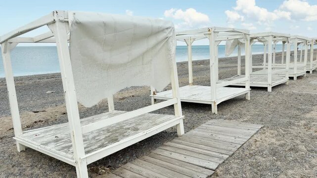 A line of worn wooden beach gazebos rests silently on the empty shore. The weathered fabric and deserted boardwalk create a peaceful yet melancholy feeling of the off-season.
