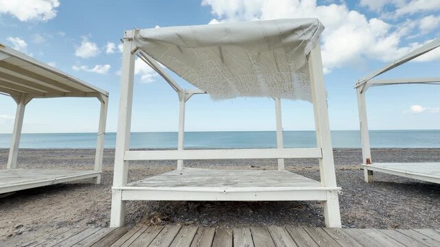 Weathered wooden cabanas face the calm blue sea under a partly cloudy sky. The shredded white fabric and empty shoreline stir feelings of nostalgia and quiet serenity.
