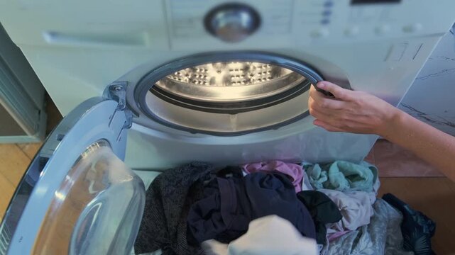 Close perspective of female hands placing mixed garments into washing machine drum, adjusting load before starting cycle, everyday home laundry process and fabric care routine indoors.