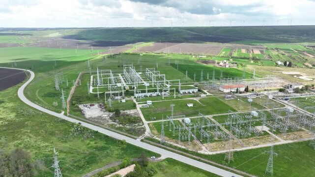 Aerial view of an electrical substation and high-voltage power lines with a wind farm on the horizon. Energy grid infrastructure, symbolizing the transition to renewable power.