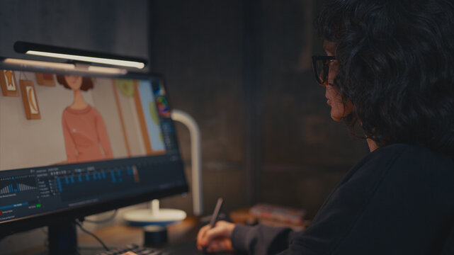 Side profile shot of female artist with curly hair and glasses, drawing on graphic tablet in warm loft studio. Focus on artist looking at monitor with cartoon character illustration in progress.