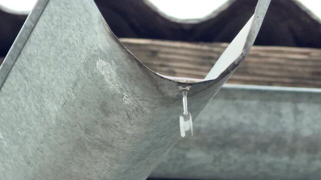A single water drop drips from the edge of a weathered metal gutter. The quiet and peaceful moment captures a calm, gray rainy day in the neighborhood.