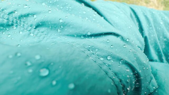 Shiny raindrops bead up beautifully on a vibrant teal waterproof jacket. The fresh droplets sit perfectly on the protective fabric, creating a calm and refreshing outdoor mood.
