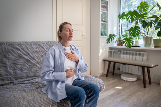 Positive mature woman do stress relief deep breathing self helping therapy, female sitting on sofa holding hands on chest inhaling air, focusing on physical and mental relaxation, healing zen practice