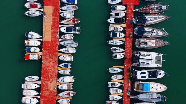 Top view aerial of luxury yachts docked at red pier. Drone shot looking down at rows of white boats moored at a harbor pier.