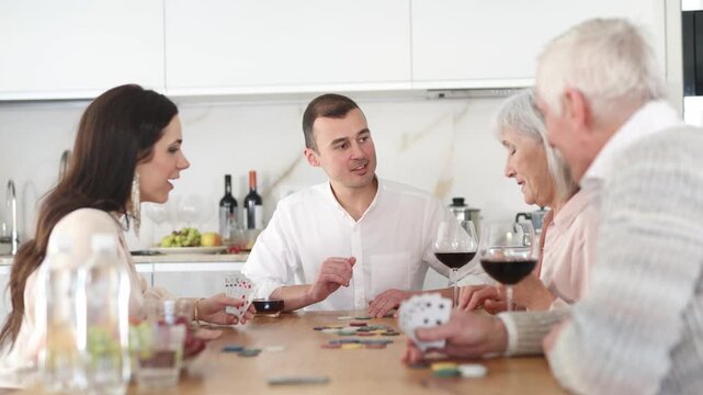 Joyful senior parents and young couple sitting around table playing with cards and drinking wine in goblets at home