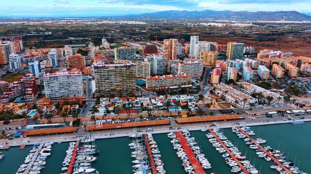 Aerial view of luxury yachts moored in Spanish marina port. Wide drone shot of many white boats docked at red piers in turquoise sea water with city background.