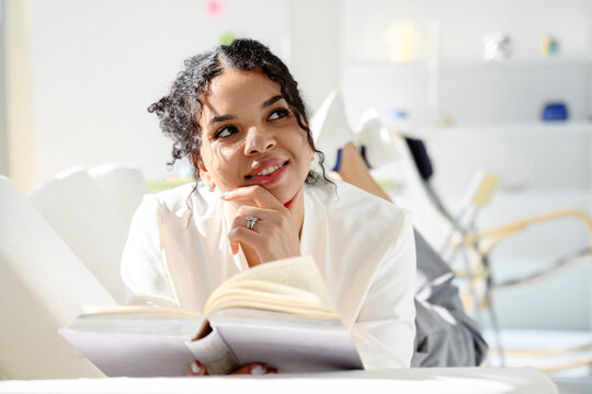 Woman lying on sofa indoors reading a book and daydreaming