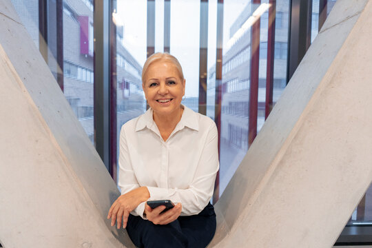 Happy senior businesswoman sitting with smart phone on columns in office
