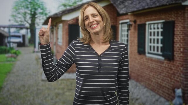 Woman pointing index finger and smiling beside a brick building on a cobblestone path with shuttered windows and garden; joyful.