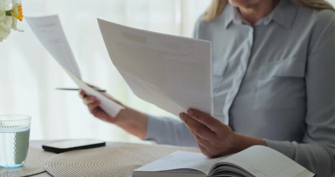 Unrecognisable elderly lady looking through utility bills putting them on round coffee table and typing on laptop.