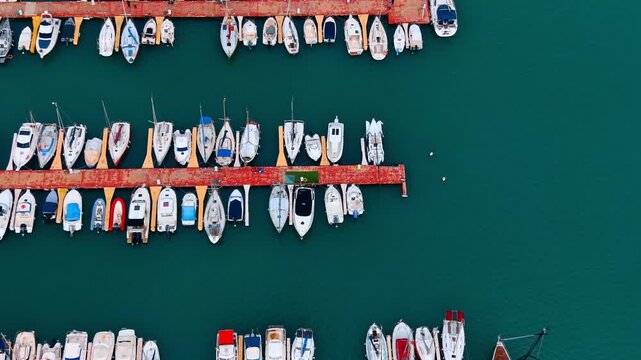 High angle vertical shot of sailing yachts at pier. Detailed top view of various white boats organized along a red dock in calm turquoise water.