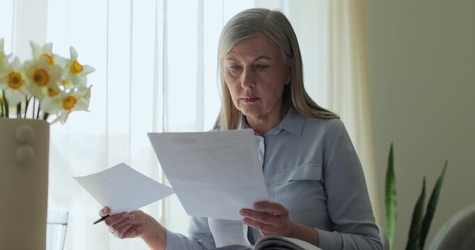 Close up older gray-haired woman sits at a table in the living room, calculating bills and taxes and taking notes.