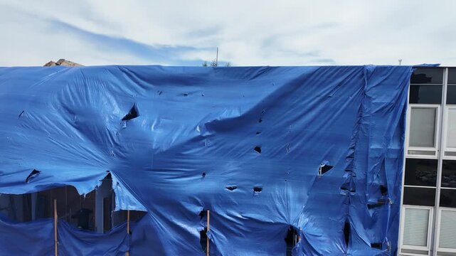 Tattered blue tarp billows in the wind against a cloudy sky and old building. A large, torn blue tarp is dramatically illuminated by sunlight, flapping in the wind, revealing glimpses of an old