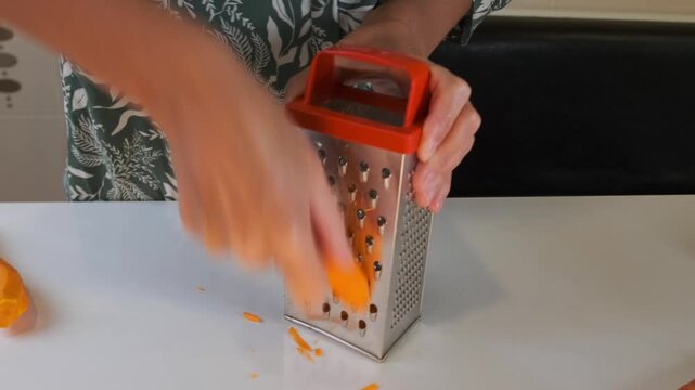 Woman grating fresh carrot on metal grater, front view of shredding vegetable for cooking, hands preparing ingredient on kitchen surface, healthy meal preparation process at home