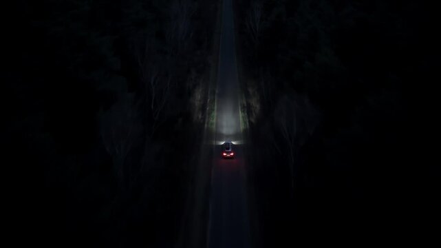 Car driving along straight road in dense forest at night filmed from drone with tracking movement and visible headlights