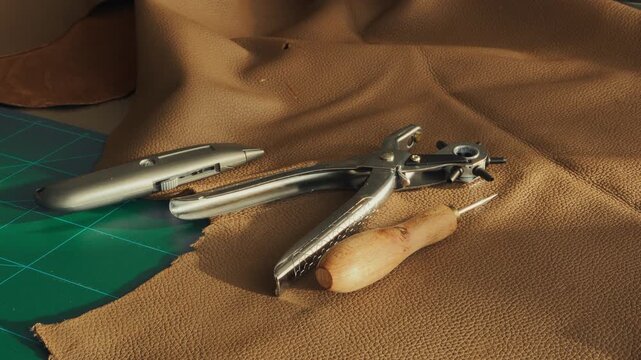 Handheld Close-up of leathercraft tools on a workbench in an artisan workshop, including a hole punch, utility knife, and textured leather material on a green cutting mat. Warm evening light.