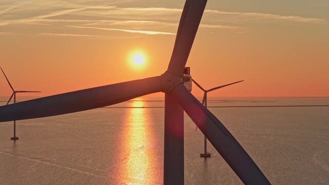 Close up aerial view of a wind turbine spinning in an offshore wind farm. The sun sets over the ocean, creating a beautiful orange sky