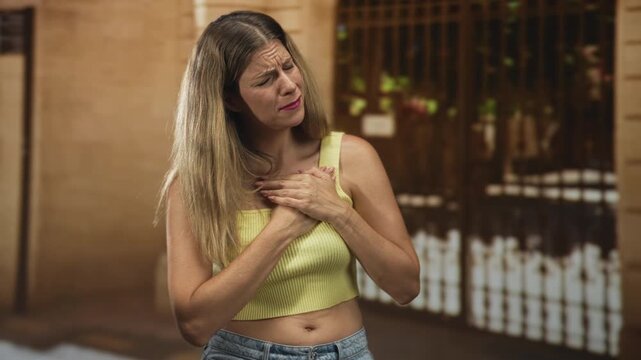 Woman clutching her chest with both hands on a street in front of a building facade wearing a yellow crop top and jeans; emotional distress vulnerability.