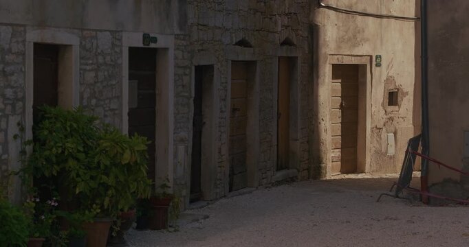 A narrow stone street shows several wooden doors, potted plants, and sunlight falling on building facades.