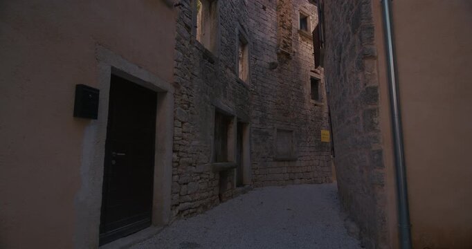 Shot of old stone buildings and narrow passage in Bale, Istria, Croatia. The scene shows stone walls, windows, shutters, doors, gravel ground and a drainpipe between closely spaced buildings.
