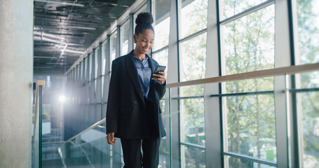 Fototapeta premium Young Businesswoman Walks Through a Bright Glass Office Hallway, Checking Her Smartphone to Track Sales Pipeline, Log Client Updates, Adjust Revenue Forecasts, and Coordinate Cross-functional Tasks