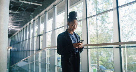 Fototapeta premium Young Businesswoman Checks Her Smartphone in a Bright Glass Office Corridor, Reviewing Project Timelines, Confirming Meetings, and Sending Quick Status Updates to Keep Team Communication