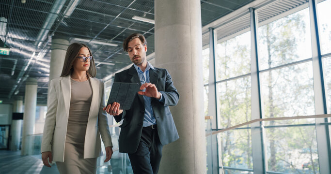 Two Focused Business Professionals Walk Through a Bright Office Atrium Reviewing a Digital Tablet, Checking Project Status, Adjusting Priorities, Confirming Deadlines, and Coordinating Next Actions