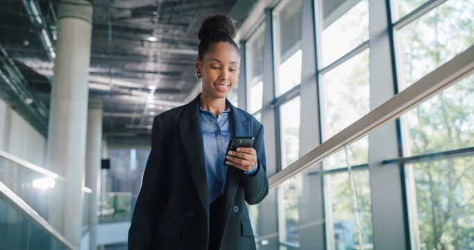 Young Businesswoman Walks Through a Bright Office Corridor, Checking Her Smartphone to Review Sales Metrics, Confirm Investor Calls, Align Team Priorities, and Capture Quick Action Items