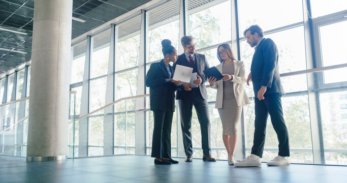 Business Team Stands in a Bright Office Hallway, Reviewing Digital Reports on Laptop and Tablet, Aligning Project Priorities, Discussing Client Updates