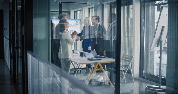 Business Team in a Glass Meeting Room Finishes a Successful Negotiation, as Two Leaders Shake Hands While Coworkers Applaud, Review Charts on a Screen, Confirm Targets, and Agree on Next Steps