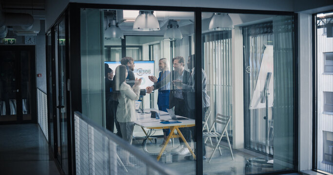 Business Team in a Glass Conference Room Celebrates Closing a Successful Deal, Shaking Hands and Applauding Around a Table With Laptops, Digital Charts, and Documents, Confirming New Partnership Goal