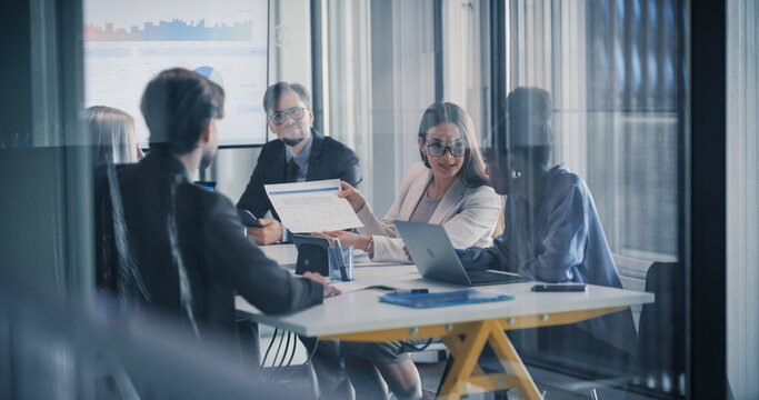 Business Team Sits in a Glass Meeting Room While a Manager Presents Printed Performance Data, Leading a Focused Review of Charts, Digital Dashboards, and Project Results to Align Goals, Budget