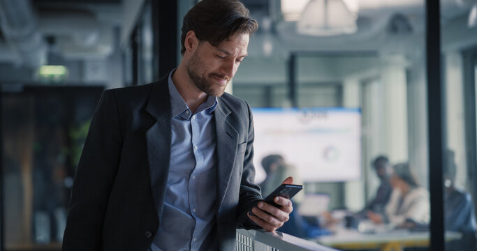 Business Executive Checks a Strategic Message on His Smartphone While Walking Through a Modern Office Corridor, Coordinating Quick Decisions as a Project Team Reviews Reports, Performance Charts