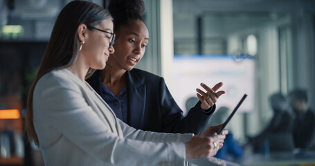 Female Business Professionals Stand in a Modern Office Hallway Reviewing a Digital Tablet, Updating Sales Pipelines, Refining Customer Journeys, Planning Product Launches, and Aligning Revenue Target. © Gorodenkoff