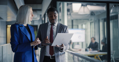 Two Coworkers Stand in a Glass Office Hallway Reviewing Sales Dashboards on a Laptop, Aligning Revenue Targets, Planning Client Follow-up Tasks, and Coordinating Next Steps While a Strategy Workshop 