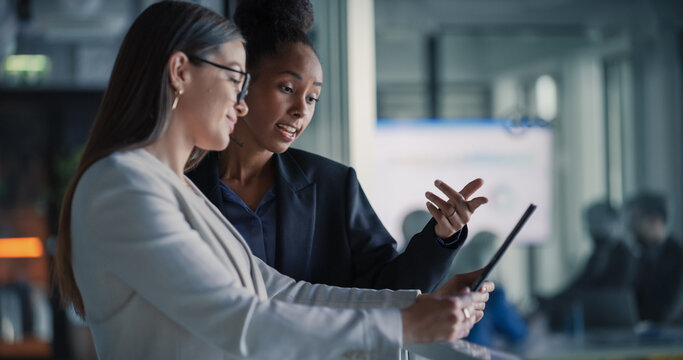 Female Business Professionals Stand in a Modern Office Hallway Reviewing a Digital Tablet, Updating Sales Pipelines, Refining Customer Journeys, Planning Product Launches, and Aligning Revenue Target.
