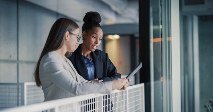 Two Businesswomen Stand in a Modern Office Hallway Reviewing a Digital Tablet, Discussing Project Updates, Clarifying Priorities, and Planning Next Steps to Improve Client Service, Team Coordination