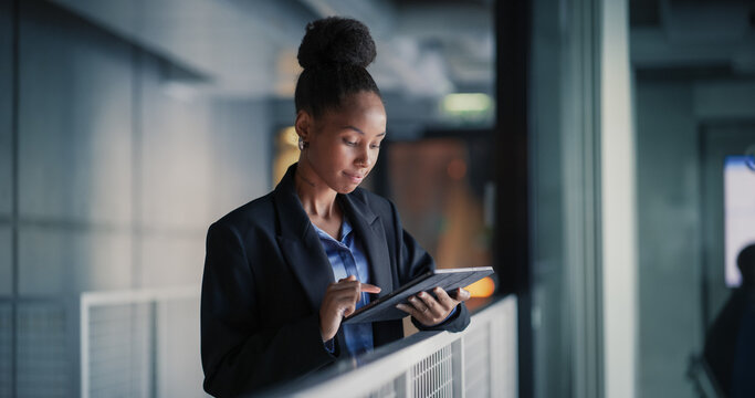 Focused Young Businesswoman Stands by an Office Railing Using a Digital Tablet to Check Sales Metrics, Update Client Lists, Organize Tasks, and Adjust Project Timelines During a Quiet Evening Work 