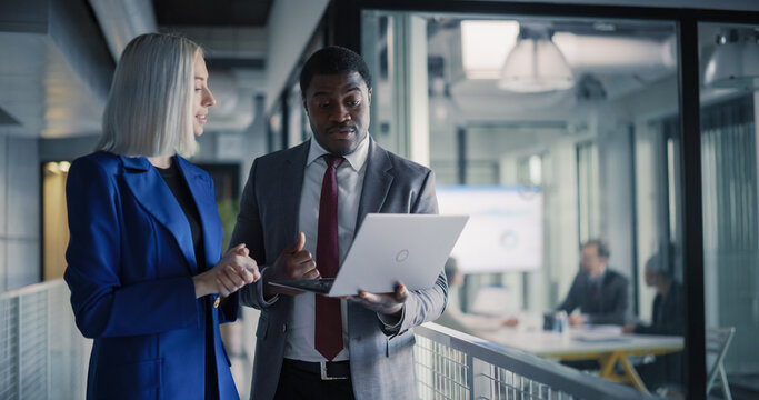 Two Coworkers Stand in a Glass Office Hallway Reviewing Sales Dashboards on a Laptop, Aligning Revenue Targets, Planning Client Follow-up Tasks, and Coordinating Next Steps While a Strategy Workshop 