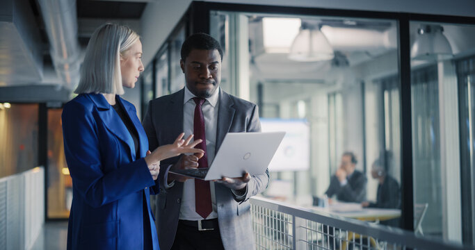 Coworkers Review Project Metrics on a Laptop in a Modern Office Hallway, Align Priorities, Clarify Tasks, and Plan Next Actions While a Business Meeting Continues in a Glass Conference Room