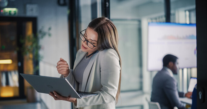 Business Professionals Work in a Modern Office Hallway, as a Focused Woman Balances a Client Call With Laptop Analysis While a Colleague Studies Performance Dashboards on a Large Screen, Coordinating