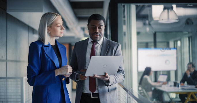 Two Coworkers Stand in a Modern Office Corridor Reviewing Performance Dashboards on a Laptop, Aligning Revenue Targets, Refining Client Strategy, and Coordinating Cross-functional Initiatives