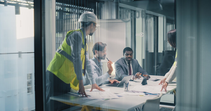 Executives and Engineers Meet in a Glass Conference Room to Review Construction Plans, Align Budget and Schedule, Track Progress on Laptops, and Coordinate Risk Control, Resource Allocation