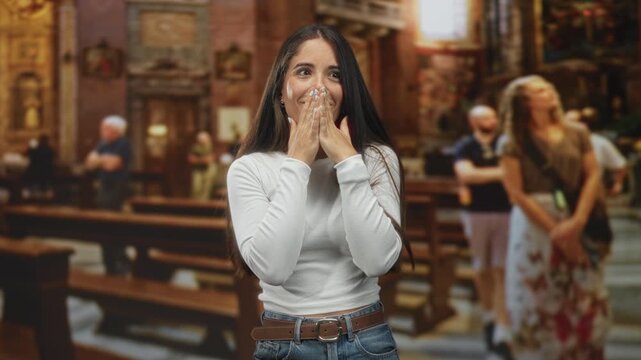Woman covers mouth with hands inside ornate church building by wooden pews and paintings with tourists; awe surprise.