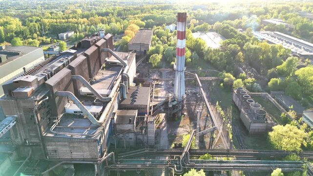 Industrial manufacturing plant with smokestack and extensive piping system surrounded by lush greenery, showcasing the facility's structure and operational elements in a vibrant environment. Aerial vi