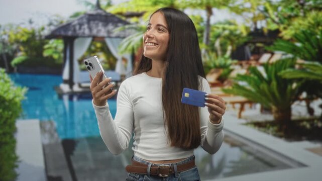 Woman holding creditcard and smartphone, smiling while posing in a studio pool set with cabana and palms; travel planning confidence.