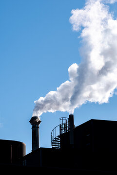 Industrial chimney and steam plume above a roof plant in blue sky with silhouette contrast defining a clean graphic stack composition at daylight