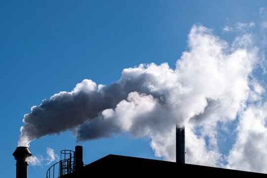 Industrial chimney and smoke plume above a roof plant in blue sky with silhouette contrast forming a graphic stack against daylight